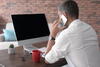 a man in a white shirt at his computer desk making a call on his mobile phone